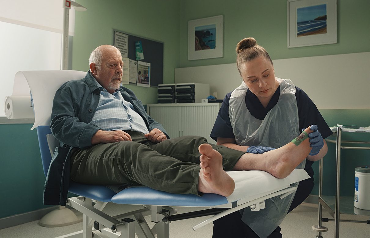 Vårdpersonal i vårdmiljö som applicerar ett Sorbact-förband på en äldre mans fot.